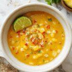 A vibrant bowl of Mexican Street Corn Soup featuring bright yellow corn, garnished with fresh cilantro, crumbled cheese, and a drizzle of spicy sauce, served in a rustic white bowl on a wooden table with colorful toppings around.