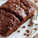 A sliced piece of moist chocolate pumpkin bread on a rustic wooden plate, revealing rich, dark chocolate swirls and vibrant orange pumpkin texture, garnished with a light dusting of powdered sugar. The bread's surface is moist and tender, with a slightly crumbly crust and a rich, inviting appearance.