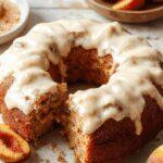 A sliced peach cake featuring a golden-brown crust and moist interior, topped with a creamy brown sugar frosting. The cake is displayed on a rustic wooden table with a few peach slices and a spoon nearby, highlighting its inviting, homemade appeal.