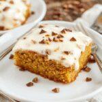 A beautifully layered pumpkin cake with rich, creamy frosting topped with sugared pecans and cinnamon, presented on a rustic wooden platter, showcasing its moist texture and festive fall decor in the background.