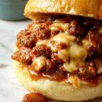 A close-up of a hearty French Onion Beef Sloppy Joe in a toasted bun topped with melted cheese and caramelized onion slices, served on a rustic plate with a side of crispy fries and fresh salad. The sandwich is overflowing with savory beef, golden-brown onions, and gooey cheese, with steam rising from the warm bread.